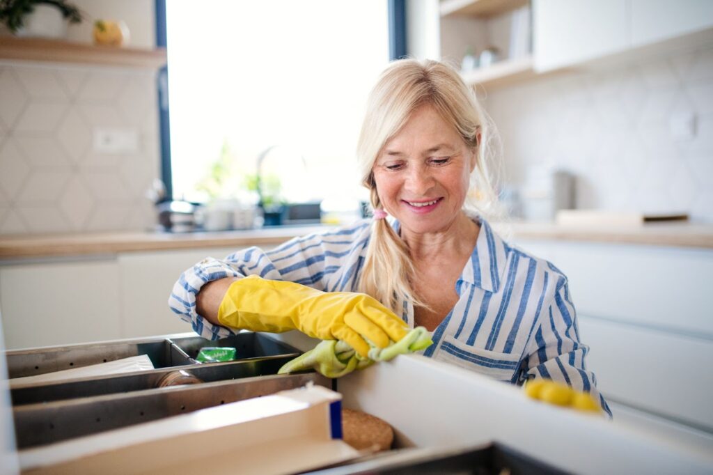 Woman smiling while cleaning kitchen drawer with yellow gloves and green cloth.