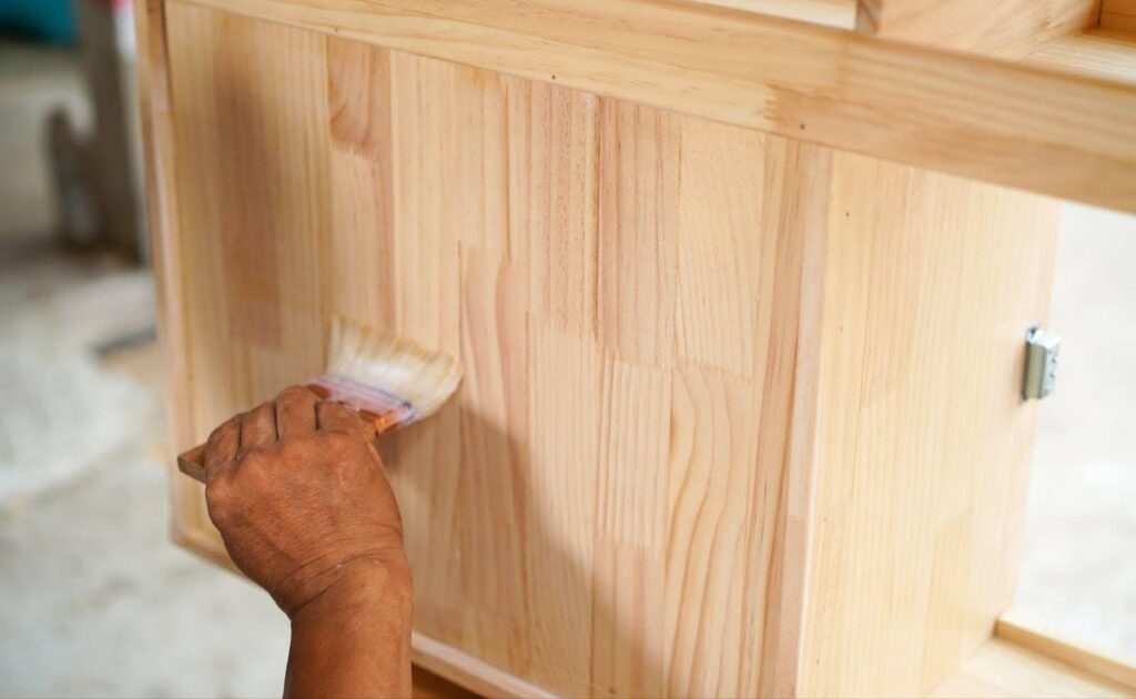 A person paints a wooden cabinet with a brush focusing on the surface to enhance its appearance