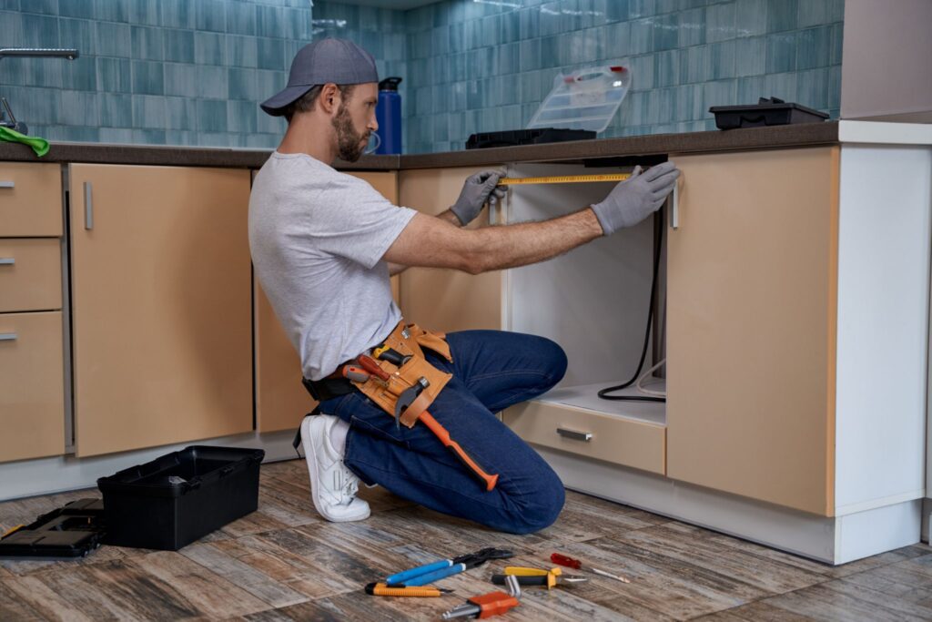 A handyman using a tape measure to measure a kitchen cabinet opening.