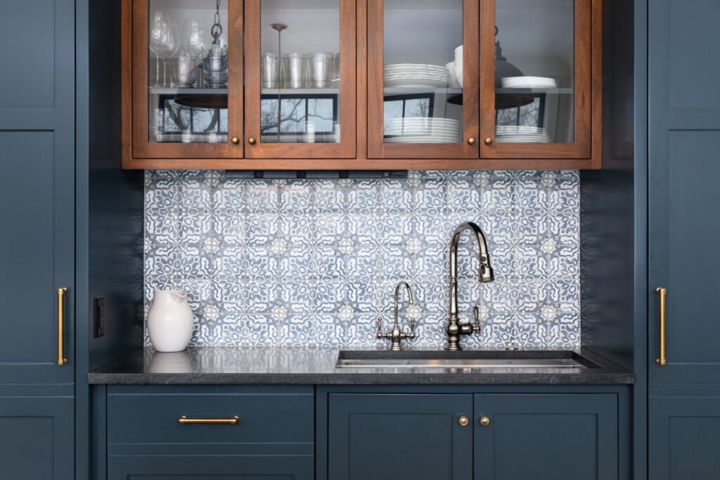 A kitchen sink area featuring dark blue lower cabinets, wood-framed glass upper cabinets, and a blue and white patterned tile backsplash.