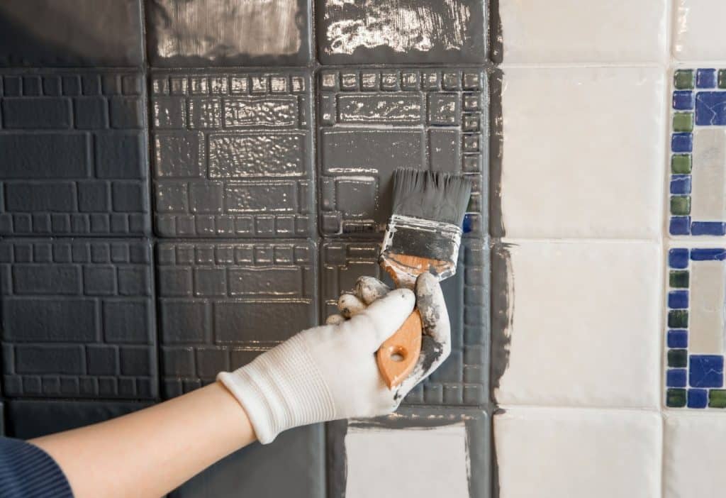 A person painting a tile wall with a brush, showcasing an affordable bathroom remodel technique to save money.