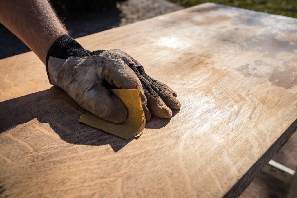 A person wearing gloves sands a wooden table, focusing on smoothing its surface for a polished finish.