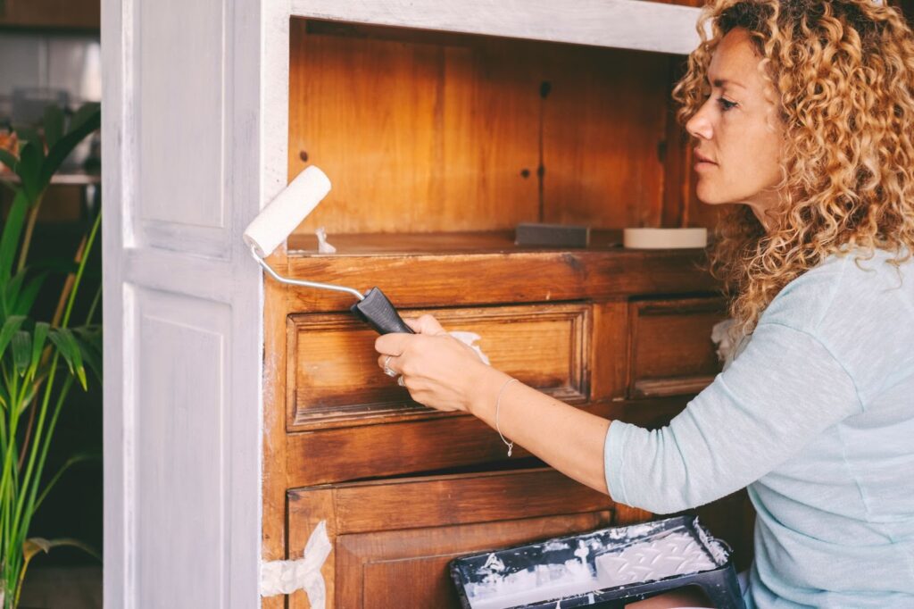 A woman is using a paint roller to apply paint to a wooden cabinet in a well-lit room.