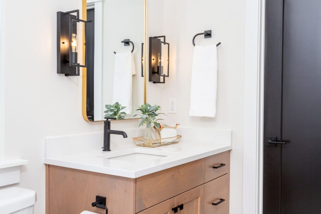 A modern bathroom vanity featuring a light wood cabinet, white countertop, black fixtures, and a gold-rimmed mirror flanked by industrial-style wall sconces.