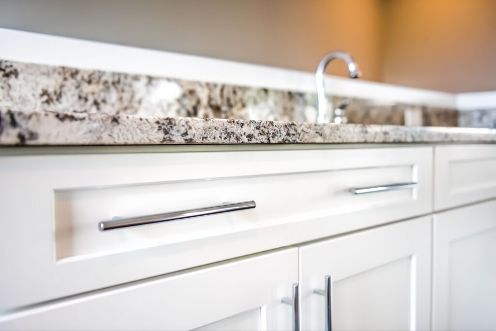 Close-up of white kitchen cabinets with chrome bar handles and a beige speckled granite countertop.